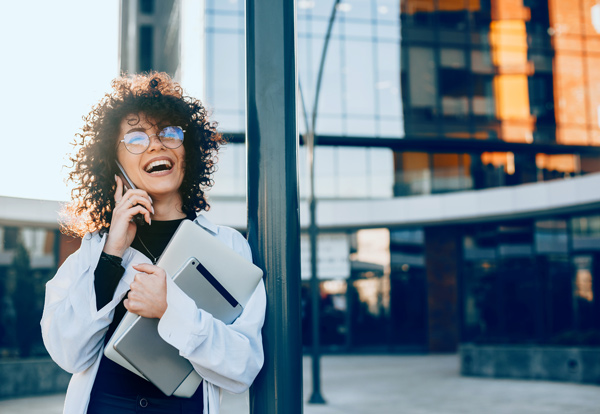 smiling woman talking on phone wearing sunglasses holding laptop in modern urban setting